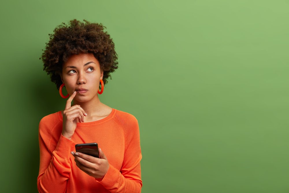Une femme qui réfléchit en tenant sont téléphone dans sa main. Elle est habillée en orange avec un fond vert.